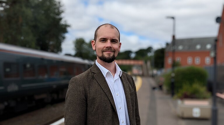 Ed, a white man with short hair and a beard, standing on the platform at Crediton station smiling at the camera